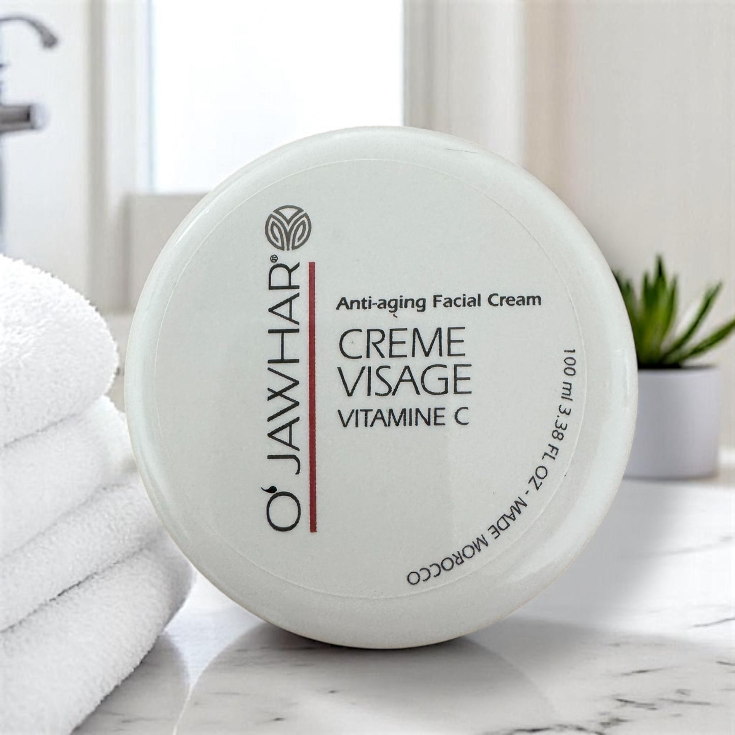 Crème visage O’Jawhar à la vitamine C – soin hydratant 100 ml dans un intérieur lumineux, posée sur une surface en marbre, à côté de serviettes blanches et d’une plante verte.