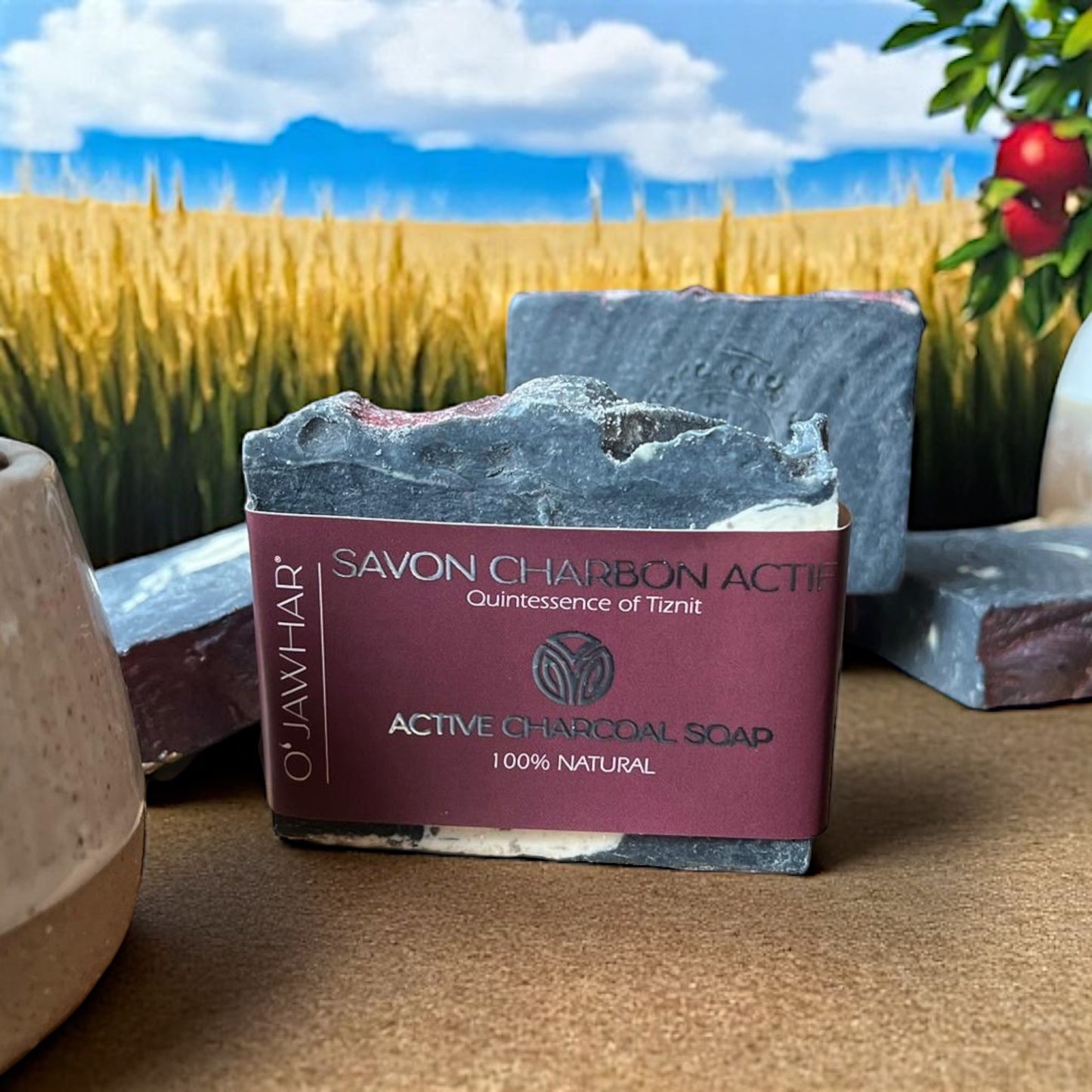 Savon Charbon Actif O’Jawhar étiqueté, posé sur table devant un champ de blé et des pommes — soin purifiant à base de charbon, inspiré de Tiznit.