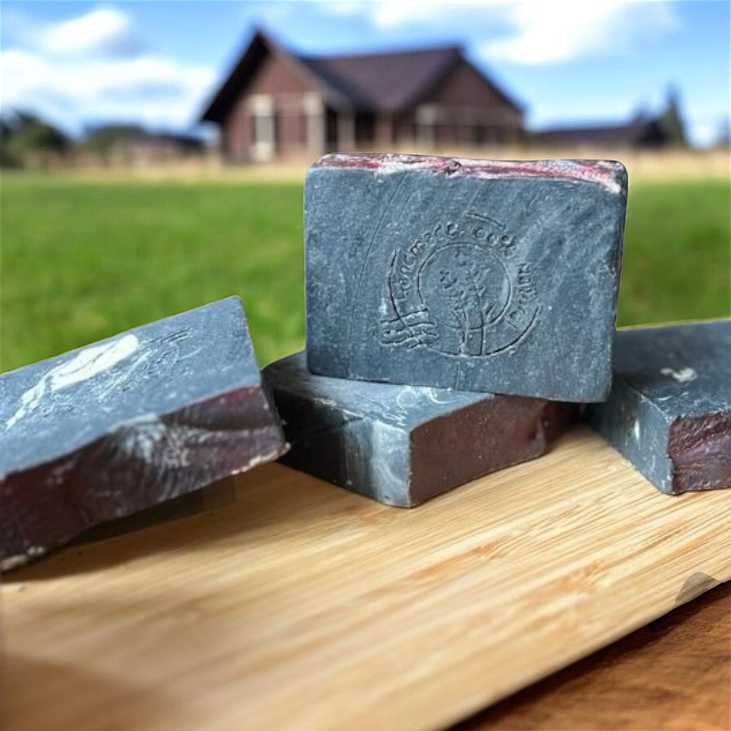 Savon au charbon actif O’Jawhar sur planche en bois, devant une maison de campagne — savon naturel purifiant fabriqué au Maroc.