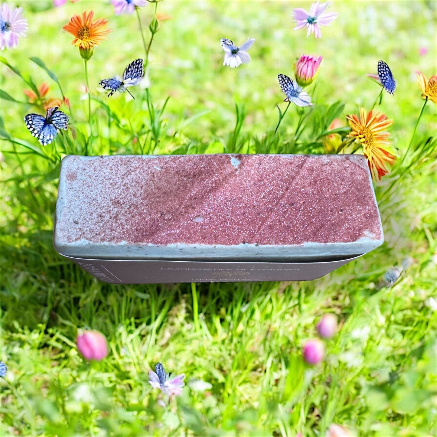 Savon exfoliant au Rhassoul O’Jawhar vu de dessus, posé dans un champ fleuri, 100% naturel et fait main