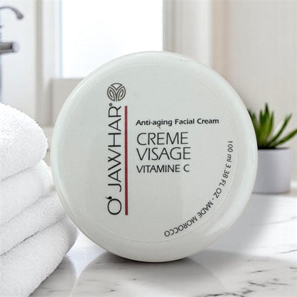 Crème visage O’Jawhar à la vitamine C – soin hydratant 100 ml dans un intérieur lumineux, posée sur une surface en marbre, à côté de serviettes blanches et d’une plante verte.